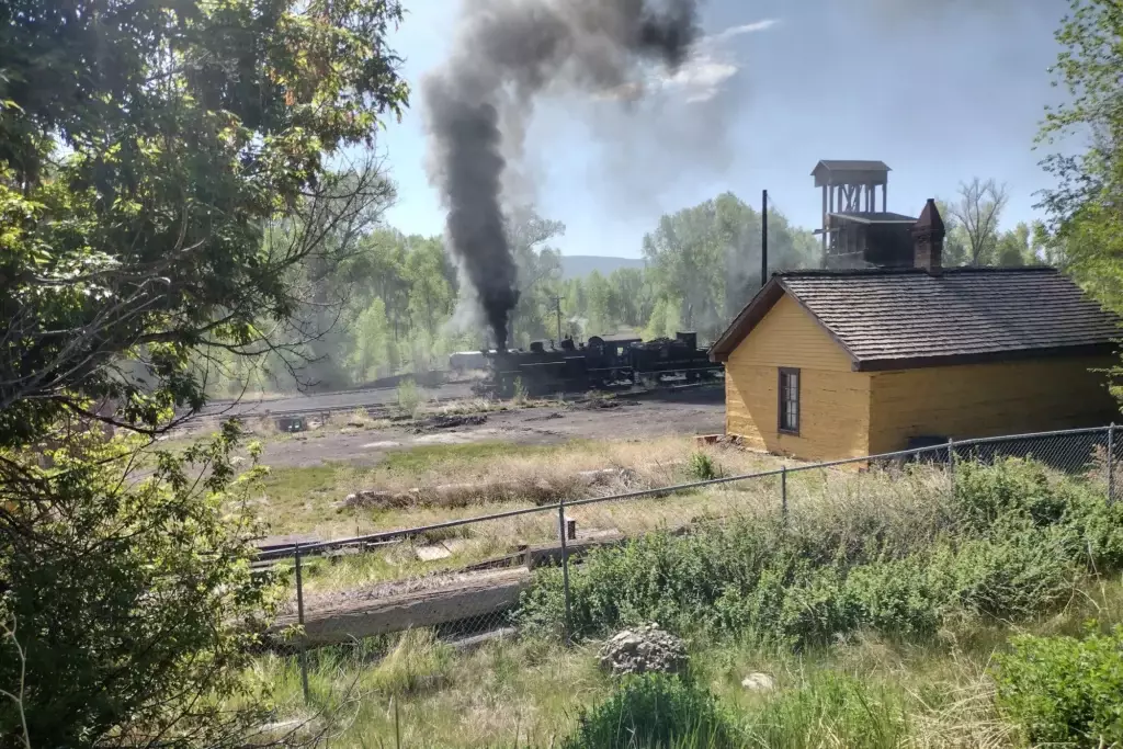 A steam locomotive emits a large plume of black smoke near a yellow wooden building in Chama, with trees and brush in the foreground, suggesting the site may once have been near a bustling hotel.