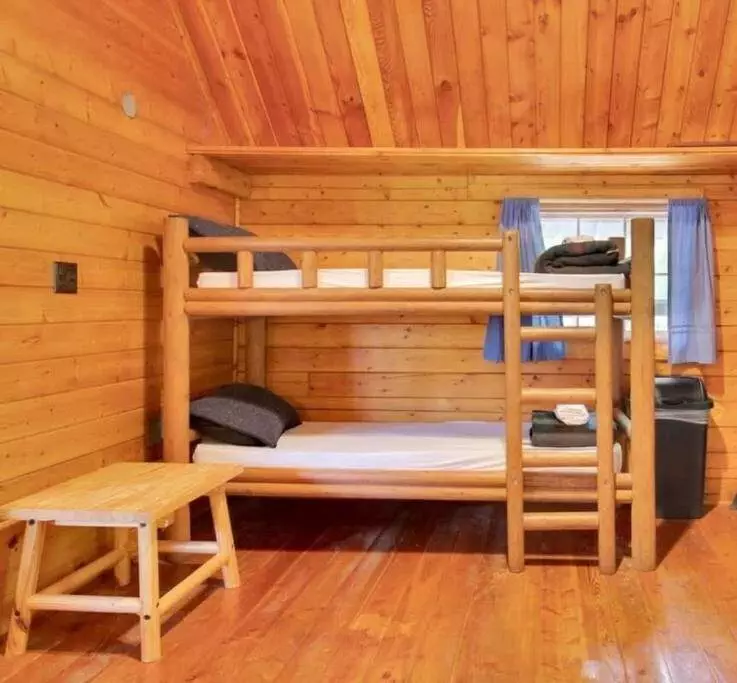 A wooden bunk bed with folded towels and pillows stands in a cozy chama hotel log cabin room with a small wooden table, blue-curtained window, and hardwood floors.