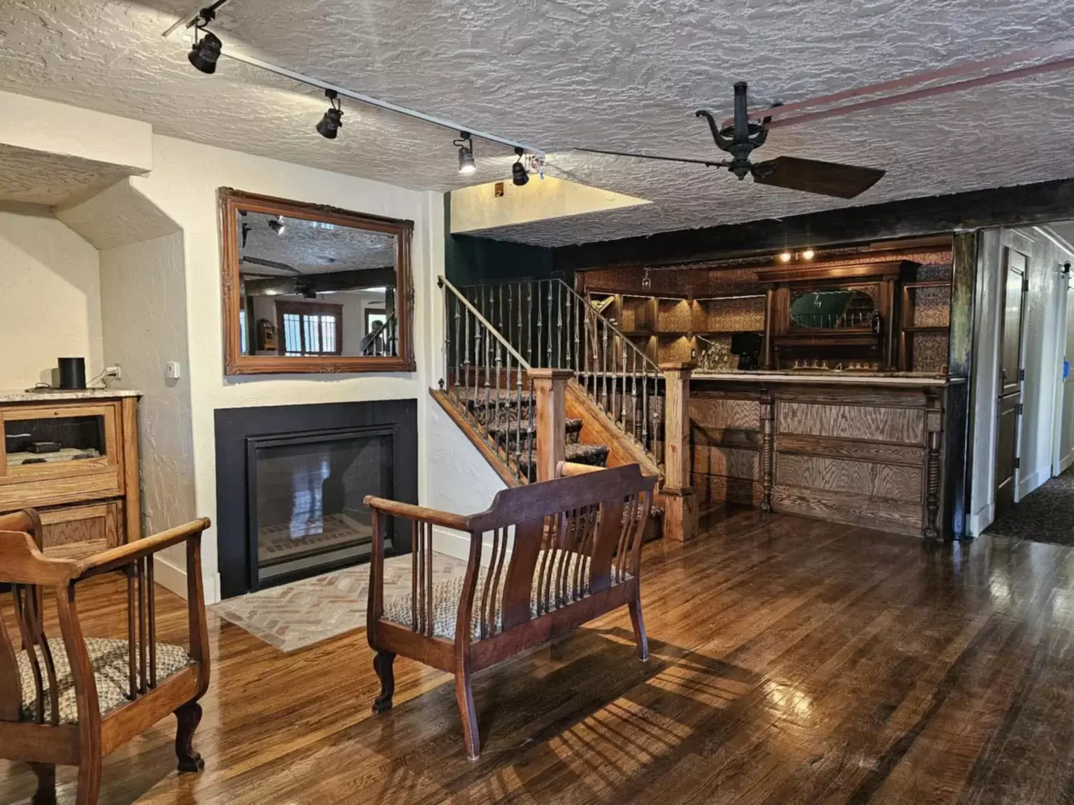 A rustic living room in Chama with wooden floors, a fireplace, a large mirror, two wood chairs, a staircase, and a built-in bar area with shelving and counter gives this space the feel of a cozy boutique hotel.