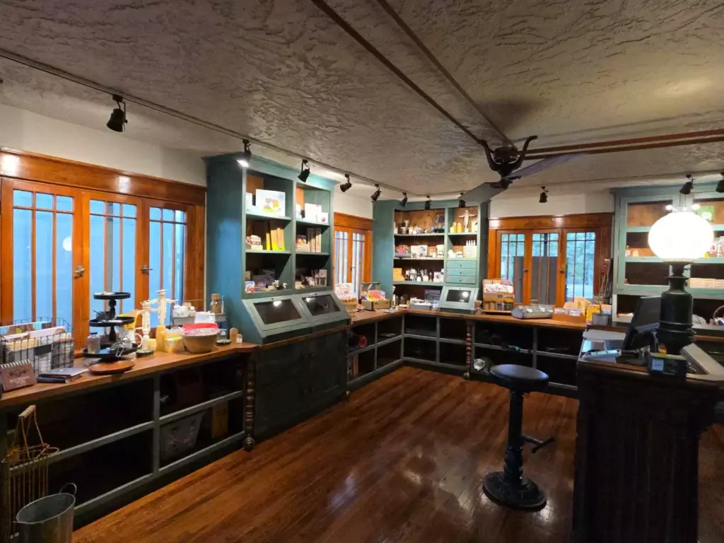 A vintage-style store interior with wooden floors, shelves stocked with assorted goods, and warm lighting evokes the charm of a historic chama hotel. An empty stool sits near the counter, inviting you to linger.
